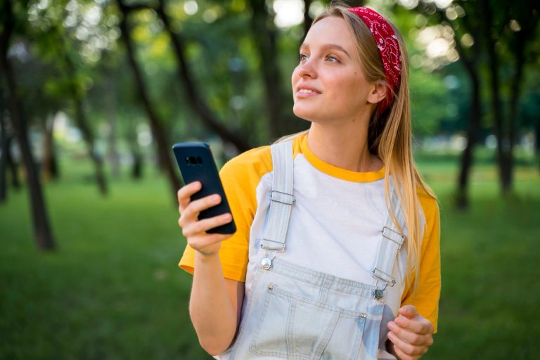 Ung kvinna med mobiltelefon i handen som tittar upp i en grön park